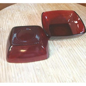 May include: Two square bowls made of deep red translucent glass. One bowl is positioned upside down, revealing a circular base with a radiating sunburst pattern, while the other bowl is upright, showing a smooth interior and rounded corners.