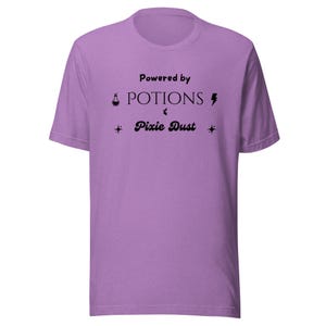 Op de afbeelding: Een lichtpaars t-shirt met korte mouwen en zwarte tekst die luidt Powered by POTIONS & Pixie Dust. Het ontwerp bevat een klein icoon van een toverdrankflesje, een bliksemschicht en twee vierpuntige sterren rondom de gedrukte tekst.