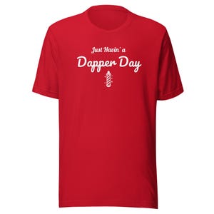 Puede incluir: Una camiseta roja de manga corta con el texto en blanco Just Havin' a Dapper Day. Debajo del texto hay un pequeño gráfico blanco de un poste de barbero clásico con rayas diagonales y pequeños detalles de estrellas en el pecho.