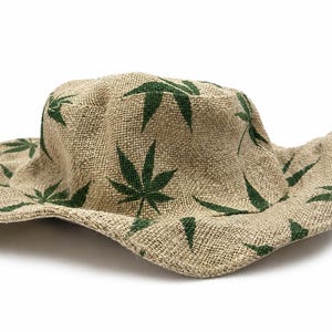 May include: A tan bucket hat made of a coarse, woven fabric featuring a repeating pattern of dark green, multi-pointed leaf shapes printed across the crown and the wide, flexible brim. The hat has a rounded top and a wavy, unstructured edge.