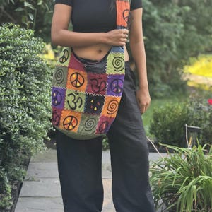 May include: A patchwork hobo bag featuring a colorful array of square fabric panels in shades of orange, green, purple, and black. The bag displays embroidered symbols including peace signs, spirals, and Om signs, with a wide shoulder strap.