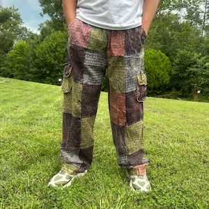 May include: Cargo pants featuring a patchwork design with various rectangular panels in shades of olive green, maroon, and dark gray. The fabric displays a repeating script pattern throughout. The pants include two side cargo pockets with flaps.