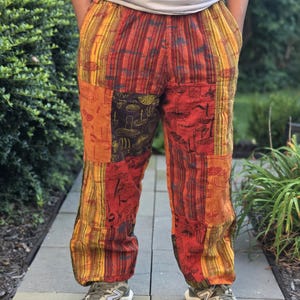 May include: Patchwork pants featuring a vibrant color palette of orange, red, yellow, and dark brown. The fabric displays a repeating mushroom print and vertical striped patterns. These loose-fitting trousers have an elastic waistband and cuffs.