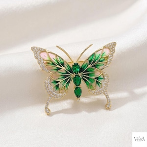 Op de afbeelding: Een goudkleurige vlinderbroche met smaragdgroene en zachtroze emaille vleugels. De vleugels zijn omlijnd met kleine, heldere pavé-gezette kristallen en het vlinderlichaam bestaat uit drie verticaal uitgelijnde, peervormige groene stenen.
