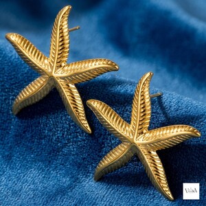 Puede incluir: Un par de pendientes de botón con forma de estrella de mar en tono dorado, con una superficie texturizada y acanalada en cada uno de los cinco brazos. Los pendientes tienen un acabado metálico y cierre de perno. Logo Virella en la esquina.