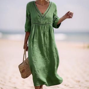 May include: A sage green midi dress featuring a V-neckline, three-quarter length sleeves, and intricate eyelet lace trim detailing along the neckline, sleeves, and waistline. The dress has a gathered skirt and is paired with a woven straw handbag.