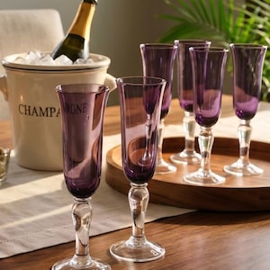 May include: A set of five tall, slender champagne flutes with deep purple glass bowls and clear glass stems and bases. The flutes are arranged on a wooden surface near a cream-colored ceramic ice bucket containing a bottle of champagne.