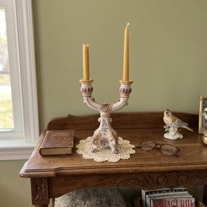 May include: A ceramic double candle holder featuring a cream-colored base with ornate floral patterns in shades of pink, green, and gold. The structure has two arms, each holding a slender, light yellow taper candle, and stands on three feet.