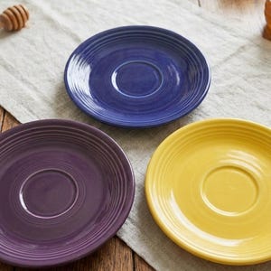 May include: Three round ceramic saucers featuring a concentric ring pattern on the rim. The set includes one deep blue saucer, one purple saucer, and one bright yellow saucer, each with a smooth, glossy finish and a central indentation for a cup.