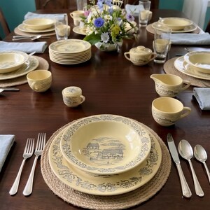May include: A dining table set with cream-colored ceramic dinnerware featuring blue landscape illustrations and floral borders. The place settings include bowls, plates, cups, a creamer, a sugar bowl, and silver flatware on woven placemats.