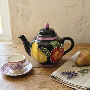 Può includere: Una teiera in ceramica nera di forma esagonale, caratterizzata da motivi dipinti di pera gialla e mela rossa, un pomello del coperchio color lavanda e un bordo della base color lavanda. Accanto si trova una tazzina bianca con piattino viola.