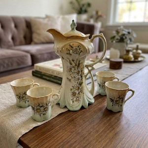 May include: A porcelain tea set featuring a tall pitcher with a lid and four matching cups. The set is cream-colored with a light green base, gold-toned rims, and a detailed floral pattern of white daisies with yellow centers on each piece.