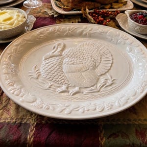Puede incluir: Una fuente de servir grande, redonda y de cerámica blanca que presenta un diseño central de pavo en relieve. El borde de la fuente está decorado con un patrón en relieve de varias frutas y follaje, creando un borde texturizado alrededor del pavo.