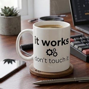 May include: A white ceramic mug featuring the black printed text "it works" above a small gear icon and the text "don't touch it" below. The mug has a standard handle and is filled with a dark brown liquid, resting on a circular wooden coaster.