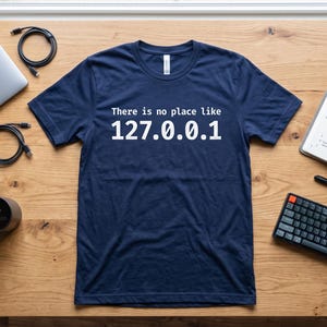 Op de afbeelding: Een donkerblauw T-shirt met korte mouwen en de witte tekst "There is no place like 127.0.0.1" op de borst gedrukt. Het shirt ligt plat op een houten ondergrond en heeft een ronde hals en een standaard pasvorm.