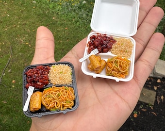 May include: Two miniature takeout containers, one black and one white, each holding a small portion of food including dark red glazed meat, light brown rice, golden brown spring rolls, and yellow noodles, accompanied by a tiny white plastic fork.