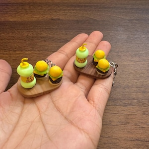 May include: Two miniature keychain sets featuring a light green Kelp Shake bottle and two small cheeseburgers arranged on a brown oval tray. Each item is crafted with detailed textures, including a yellow straw on the bottle and layered burger components, attached to a silver metal keychain ring.
