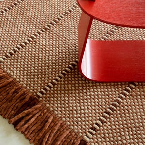 May include: A woven area rug featuring a textured grid pattern in shades of tan and brown with a thick, knotted fringe border along the edge. A smooth, curved, bright red wooden stool with a small rectangular accent piece sits on the rug.