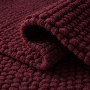 Op de afbeelding: Een diep bordeauxrood kleed met een dicht, getextureerd oppervlak bestaande uit talloze kleine, afgeronde, kiezelachtige plukjes. Het kleed is in een hoek omgeslagen, wat de dikke, consistente constructie van het lusvormige materiaal onthult.