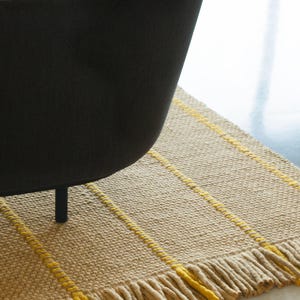 May include: A rectangular woven rug featuring a textured beige base with vertical yellow stripes and a fringed edge. The rug is partially covered by a dark gray upholstered chair with a single black leg visible on the surface of the rug.