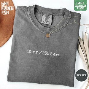 Peut inclure: Un t-shirt à manches courtes gris anthracite avec le texte brodé en blanc "in my RPSGT era" centré sur la poitrine. Le t-shirt présente une encolure ronde et une étiquette de marque Comfort Colors à l'intérieur du col, indiquant une taille S.