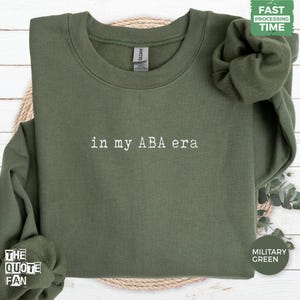 Può includere: Una felpa girocollo color verde militare con il testo ricamato in bianco "in my ABA era" centrato sul petto. Il capo presenta maniche lunghe con polsini a coste e uno scollo a coste, con un'etichetta Gildan visibile sul colletto.