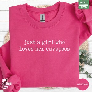 Puede incluir: Una sudadera de cuello redondo en color rosa brillante con el texto blanco "just a girl who loves her cavapoos" impreso en el pecho. La prenda tiene un cuello acanalado, mangas largas y una etiqueta de la marca Gildan en el interior.