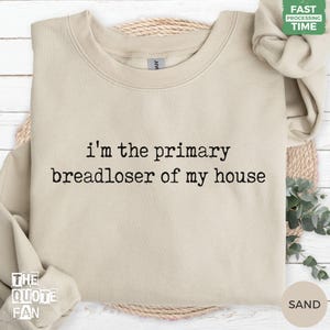 May include: A sand-colored crewneck sweatshirt featuring the black text "i'm the primary breadloser of my house" printed in a typewriter-style font across the center of the chest. The garment has long sleeves and a ribbed collar.