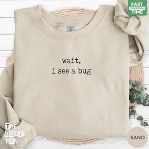 May include: A sand-coloured crewneck sweatshirt featuring black embroidered text centred on the chest that reads, wait, i see a bug. The garment has long sleeves, a ribbed collar, and a soft fabric texture, presented in a neutral tone.