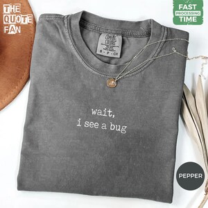 Puede incluir: Una camiseta de manga corta de color gris carbón con el texto bordado en blanco 'wait, i see a bug' centrado en el pecho. La camiseta presenta un cuello redondo y una etiqueta de la marca Comfort Colors en el interior del cuello. Una fina cadena de plata con un pequeño colgante redondo descansa sobre la tela.