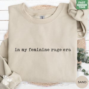 May include: A sand-colored crewneck sweatshirt featuring the black text "in my feminine rage era" printed in a typewriter-style font across the center of the chest. The garment has long sleeves, a ribbed collar, and a soft, solid-colored fabric finish.