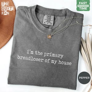May include: A pepper-colored crew neck t-shirt featuring the white text 'i'm the primary breadloser of my house' printed on the front. The shirt has a Comfort Colors brand label at the inner neckline and is paired with a thin gold-tone necklace.