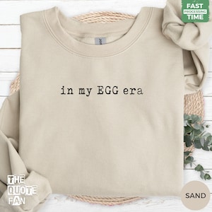 May include: A sand-coloured crewneck sweatshirt featuring the black text 'in my EGG era' printed in a typewriter-style font across the centre of the chest. The garment has long sleeves, a ribbed collar, and a soft, solid-coloured fabric construction.