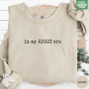 Peut inclure: Un sweat-shirt à col rond de couleur sable avec le texte brodé en noir "in my RPSGT era" centré sur la poitrine. Le vêtement présente des manches longues, un col côtelé et une coupe classique, arborant un design épuré avec une police de type machine à écrire.