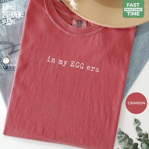 以下が含まれることがあります： 胸の中央に白い刺繍で「in my EGG era」と書かれたクリムゾンカラーの半袖Tシャツ。この衣類はリブ編みのクルーネックで、襟の内側にComfort Colorsのブランドラベルが見えています。