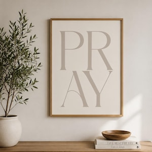 May include: A rectangular wall art print in a light wood frame featuring the word PRAY in large, light beige serif capital letters on a cream background. The print is displayed vertically, showcasing a clean, balanced composition with clear typography.