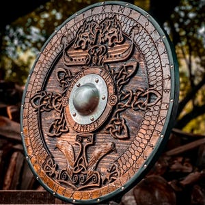 May include: A circular wooden shield featuring intricate relief carvings of two ravens at the bottom and two wolves at the top. A central metal boss is surrounded by engraved knotwork patterns and runic inscriptions along the outer rim.