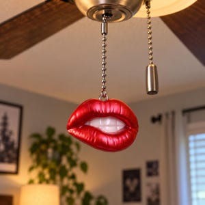 May include: A red lip-shaped ceiling fan pull chain ornament featuring a glossy finish and white teeth. The ornament is attached to a silver-toned metal ball chain that hangs from a ceiling fan fixture, which also includes a separate light pull.