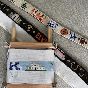 May include: Three needlepoint canvas belts featuring various embroidered motifs including a house, bottles, a basketball, a football, a pink car, sunglasses, and the text Keeneland, TCB, Elvis, and a blue letter K, mounted on a wooden frame.