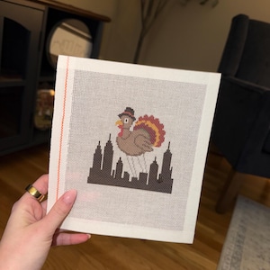 May include: A square needlepoint canvas featuring a brown turkey wearing a black top hat with a red band, positioned above a dark brown silhouette of a city skyline. The design is centered on a white grid mesh fabric with a vertical orange edge.