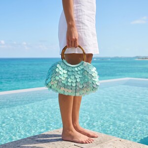 May include: A handbag covered in shimmering light blue circular sequins featuring a rounded bamboo handle. The bag has a curved, semi-circular shape and is held by a person wearing a white linen dress, showcasing the reflective surface texture.
