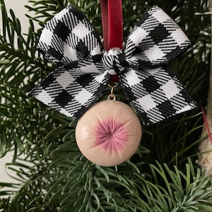 May include: A novelty Christmas tree ornament featuring a round, flesh-toned design with a pink center, suspended by a black and white checkered bow and a red ribbon. The ornament hangs among green pine needles.