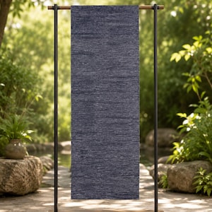 Puede incluir: Una alfombra de pasillo rectangular de color azul marino oscuro con una apariencia texturizada y jaspeada. La alfombra cuelga verticalmente sobre una barra de madera horizontal sostenida por dos postes metálicos negros sobre una base de madera.