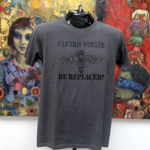 Peut inclure: Un t-shirt gris foncé avec un motif d'abeille noire et le texte "Can this worker be replaced?" imprimé sur le devant.