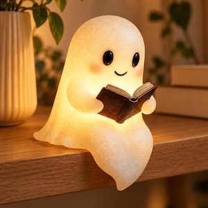 May include: A glowing, translucent white ghost-shaped lamp with a smooth, rounded form, black oval eyes, and a small smile. The ghost figure has pink-tinted cheeks and is holding a small, dark brown open book in its hands while sitting.