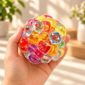 May include: A spherical sensory toy composed of numerous small, translucent, multi-colored plastic cubes in shades of red, orange, yellow, green, blue, and purple. The cubes are connected to form a flexible, textured ball with a glossy finish.
