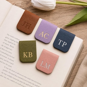 May include: Five rectangular magnetic bookmarks in brown, lavender, navy blue, olive green, and dusty pink. Each bookmark features two embossed capital letters: JH, AC, TP, KB, and LM. The bookmarks are arranged on the open pages of a book.