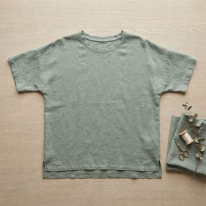 Könnte beinhalten: Ein salbeigrünes Kurzarm-T-Shirt mit Rundhalsausschnitt und geradem Saum mit Seitenschlitzen. Neben dem T-Shirt liegen ein gefaltetes Stück passender Stoff, eine kleine hölzerne Garnrolle und eine metallene Stickschere.