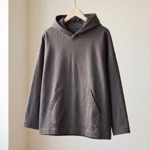 Op de afbeelding: Een donkergrijze hoodie met lange mouwen, voorzien van een kangoeroezak aan de voorzijde, een capuchon met een gestructureerde halslijn en rechte mouwen. Het kledingstuk hangt aan een houten kledinghanger en heeft een strak ontwerp.