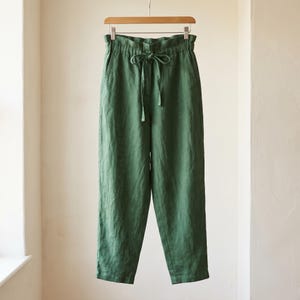 Könnte beinhalten: Waldgrüne Hose mit einer hoch taillierten, elastischen Paper-Bag-Taille und einem passenden Stoff-Bindegürtel. Die Hose hat eine lockere, gerade Passform, seitliche Eingrifftaschen und hängt an einem Holzkleiderbügel.